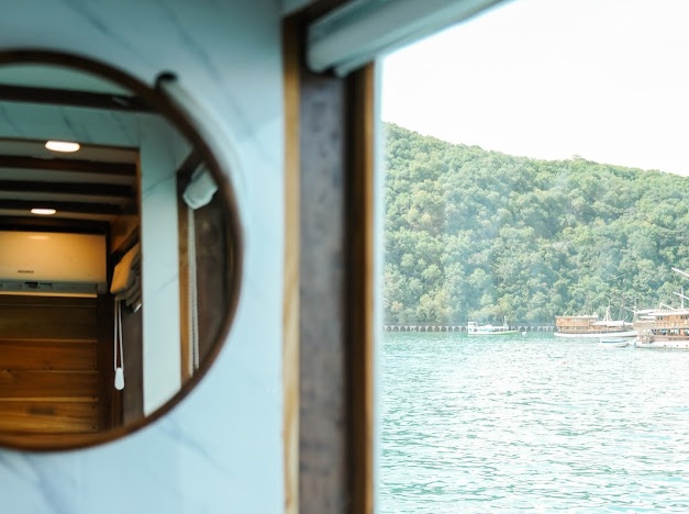 Round mirror and open window reveal green hills and anchored boats