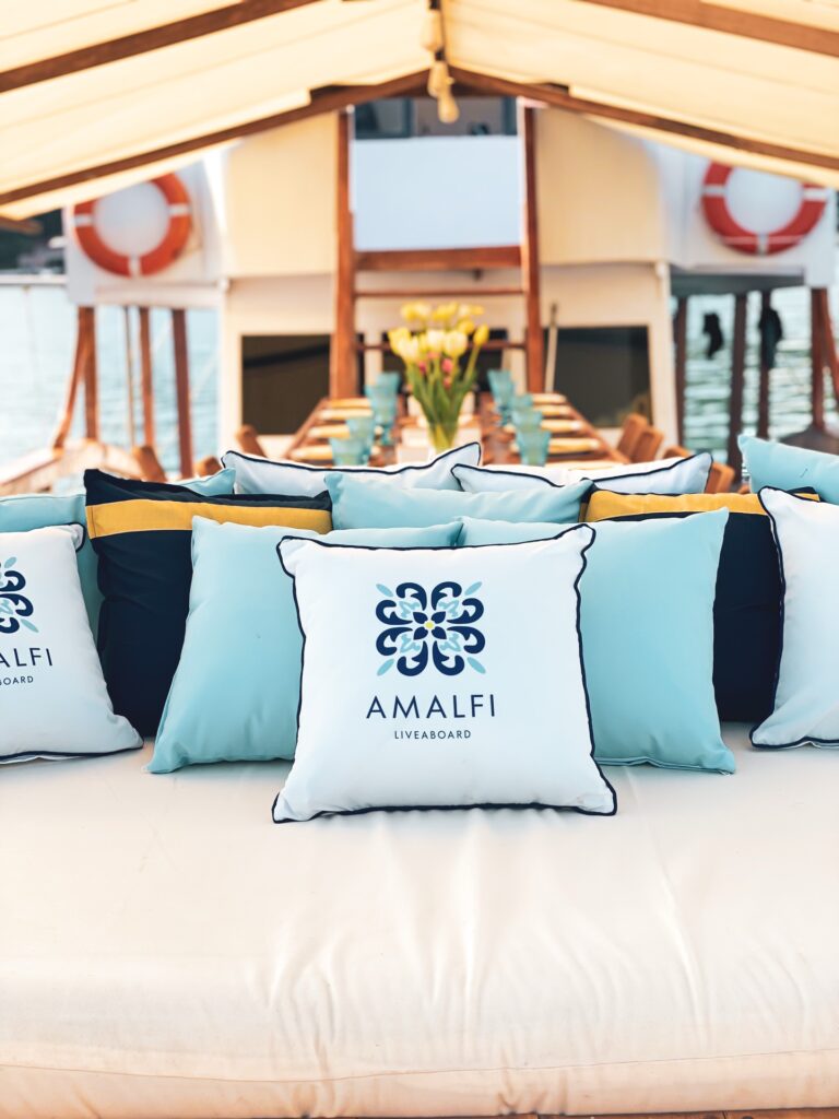 Teal and navy cushions embroidered with the Amalfi liveaboard logo arranged on a shaded deck seating bench