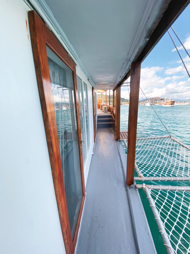 Side deck walkway with green netting rails and wood-trimmed windows overlooking the harbor
