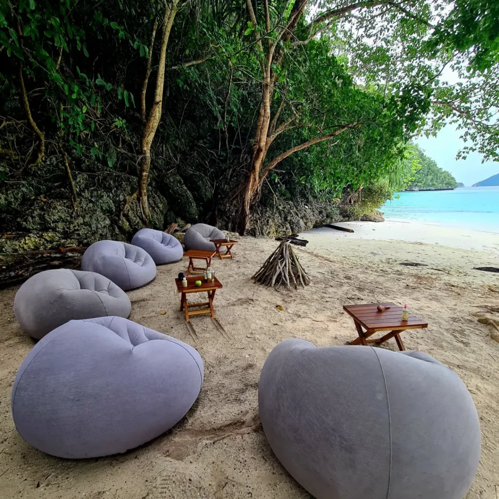 Elegant beach lounge seating arranged beneath lush jungle cliffs with plush bean bags, wooden side tables, and soft white sand beside turquoise tropical waters in Komodo Islands.