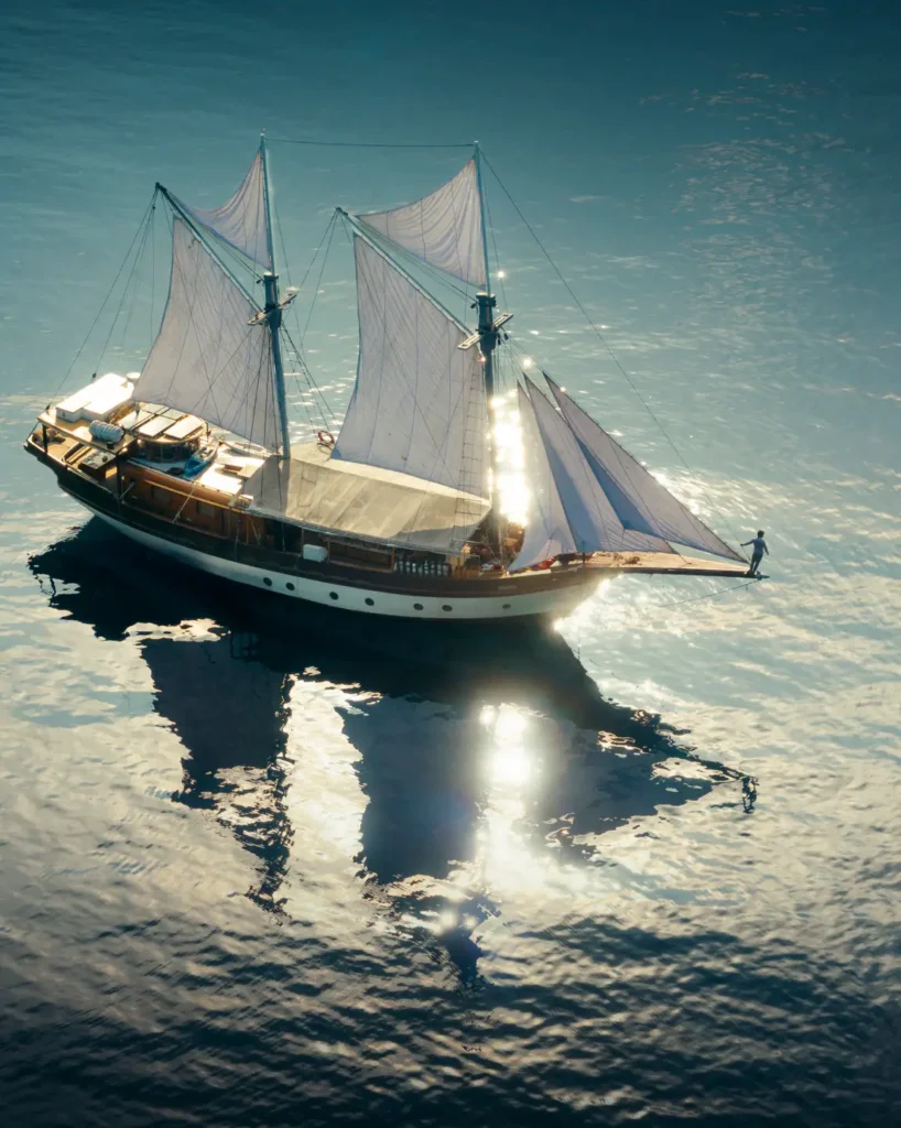 Aerial drone photograph of the Anne Bonny luxury liveaboard gliding across calm reflective ocean waters under bright tropical sunlight in Raja Ampat.