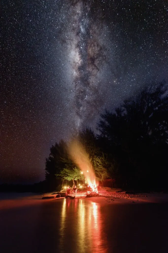 A magical beachfront bonfire experience hosted by Anne Bonny beneath the Milky Way, with glowing firelight reflecting across the shoreline and silhouetted tropical trees in Indonesia.