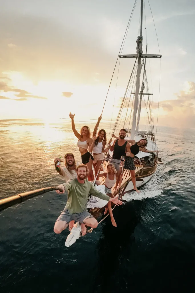 Guests celebrating together on the bow of Anne Bonny during golden hour, capturing a lively sunset moment above deep blue ocean waters with warm tropical light reflecting across the deck.