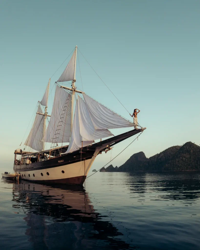 Elegant portrait of the Anne Bonny luxury Phinisi anchored in calm tropical waters with a guest standing dramatically on the bow at sunset in Raja Ampat.