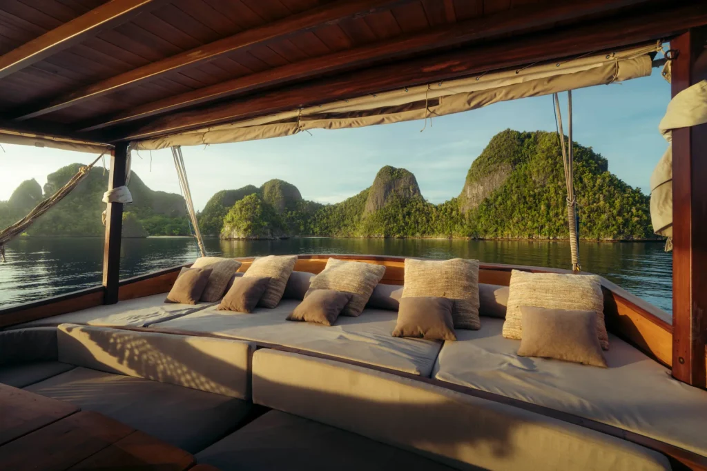Cozy open-air lounge aboard the Anne Bonny luxury Phinisi overlooking the calm limestone islands and emerald waters of Raja Ampat at golden hour.