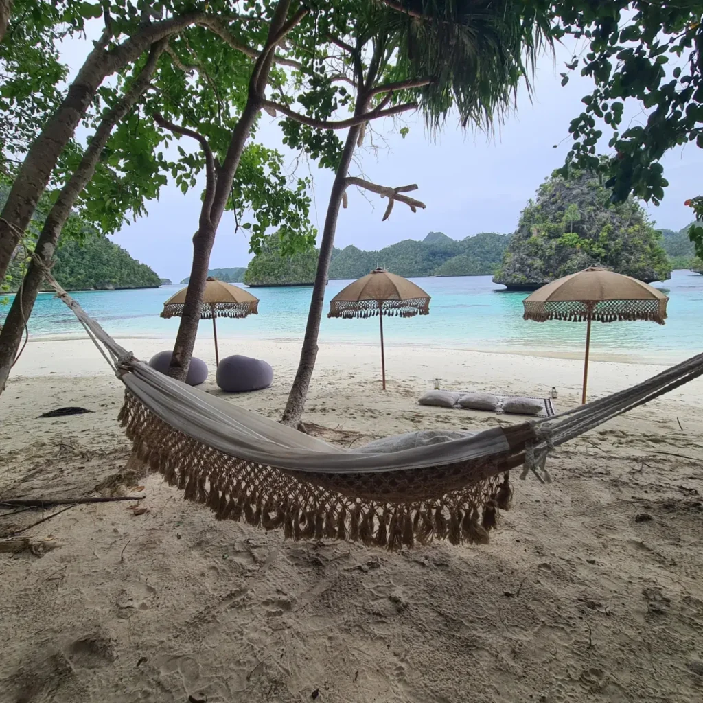 A tranquil private beach setup curated by Anne Bonny featuring handcrafted parasols, oversized lounge cushions, and a woven hammock beneath tropical trees overlooking crystal-clear water in Raja Ampat.