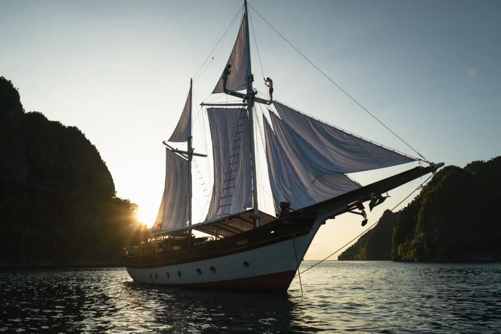 Anne Bonny anchored between towering limestone islands at sunset with its elegant white sails illuminated by warm golden light across calm tropical waters in Raja Ampat.