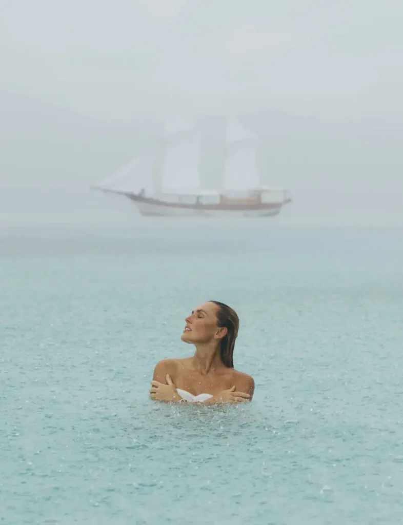 A guest swimming peacefully in calm turquoise waters while the Anne Bonny Phinisi sails softly through a misty tropical rain backdrop in Padar Island.