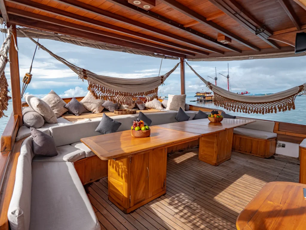 The shaded upper lounge deck aboard Anne Bonny featuring polished teak wood interiors, woven hammocks, plush wraparound seating, and open ocean views designed for relaxed luxury cruising.