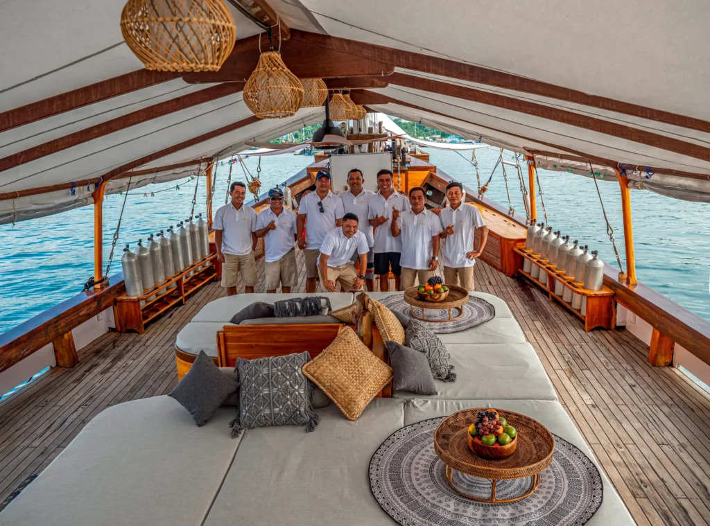 The professional Anne Bonny crew posing on the spacious shaded deck lounge surrounded by teak wood finishes, woven pendant lights, and premium liveaboard amenities ready for luxury voyages.