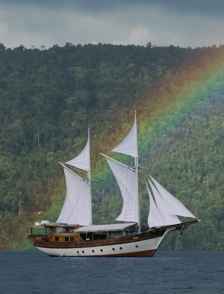 Side profile of the Anne Bonny luxury Phinisi sailing beneath a soft tropical rainbow with pristine white sails and lush rainforest islands in the background.