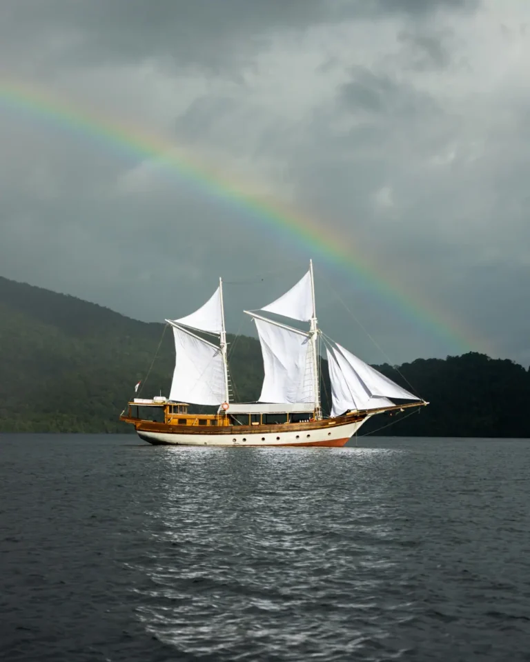 Anne Bonny Liveaboard sailing beneath a vibrant tropical rainbow with dramatic storm clouds and calm ocean waters in Raja Ampat.