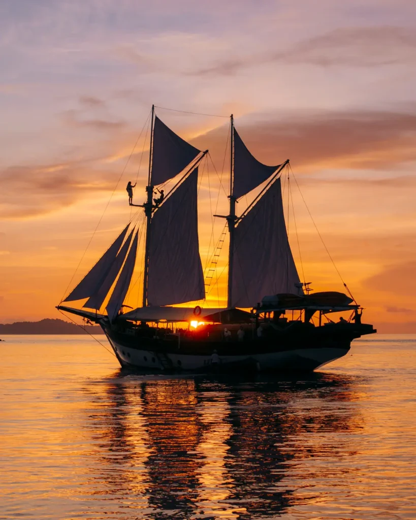 Silhouette of the Anne Bonny luxury Phinisi at golden sunset with towering sails and warm reflections shimmering across calm tropical waters.