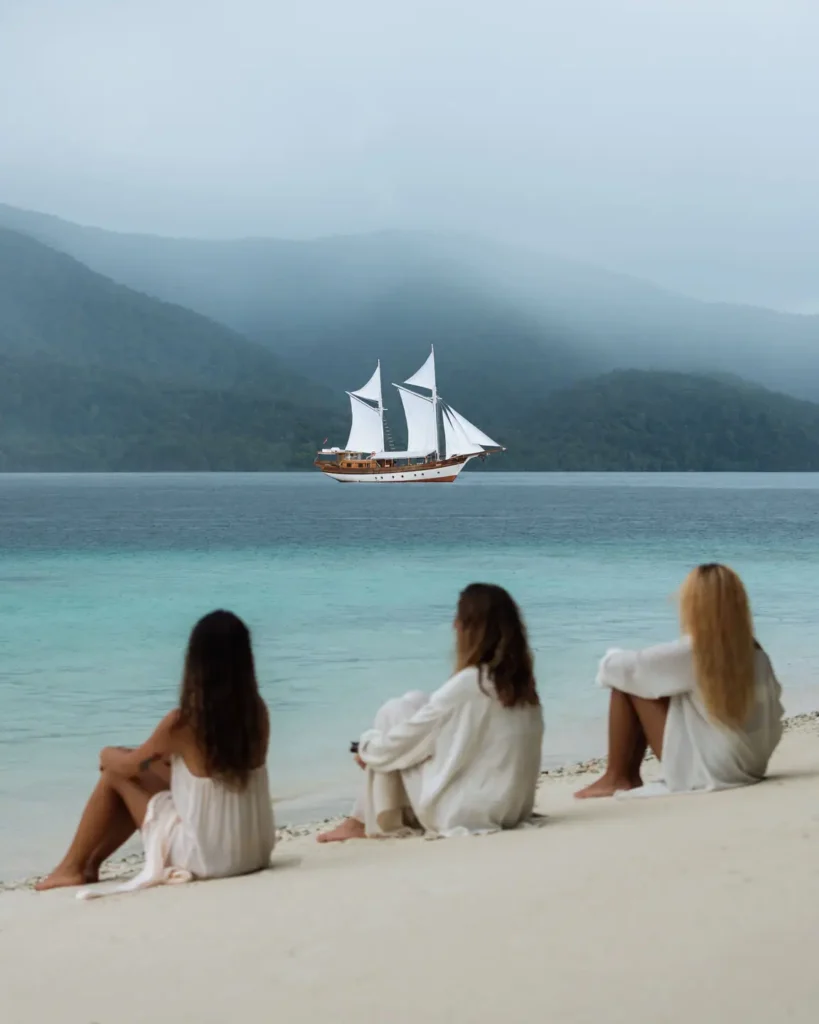 Three guests dressed in flowing white resortwear relax on a pristine white sand beach while the Anne Bonny luxury Phinisi sails across calm turquoise waters surrounded by misty jungle-covered islands in Raja Ampat.