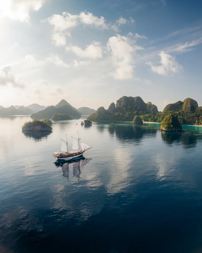 Wide cinematic aerial view of the Anne Bonny sailing peacefully through the turquoise limestone island landscapes of Raja Ampat during a private charter expedition.