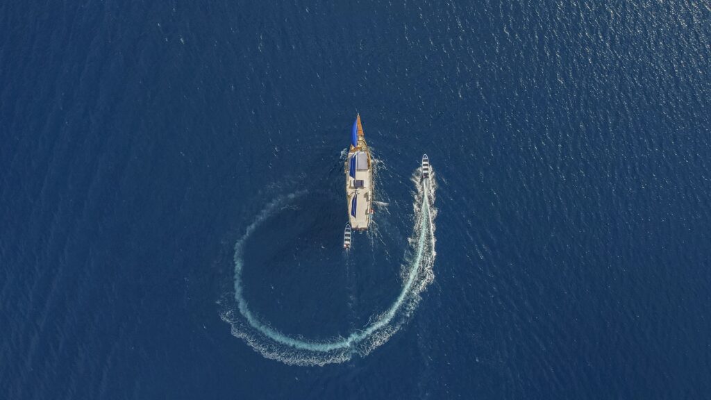 Direct top-down aerial view of Cajoma V boat tour cruising through rich blue waters while support tenders leave curved white trails behind the vessel.