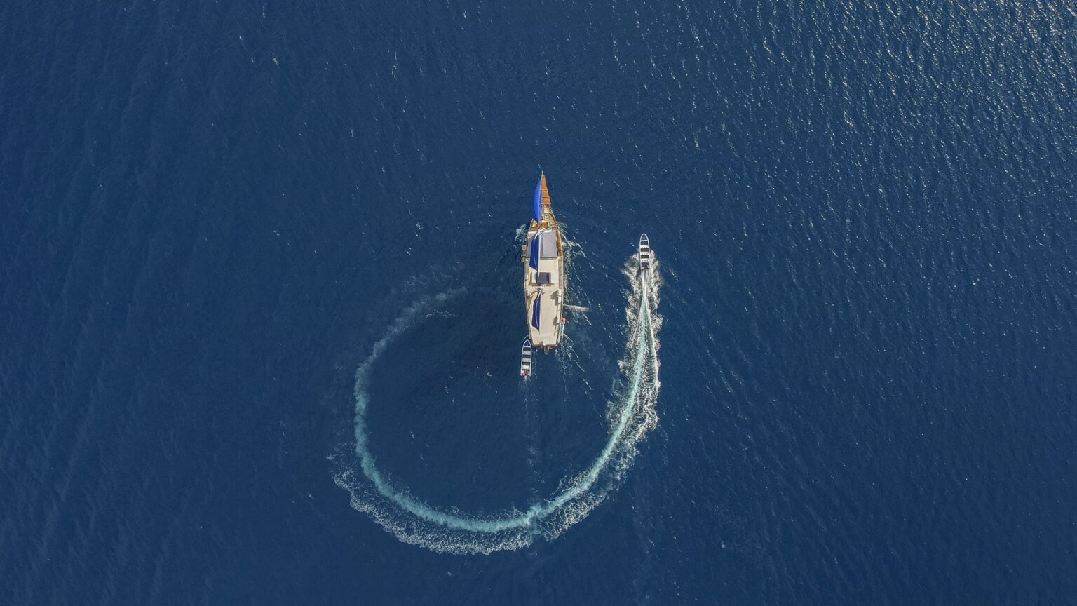 Direct top-down aerial view of Cajoma V boat tour cruising through rich blue waters while support tenders leave curved white trails behind the vessel.