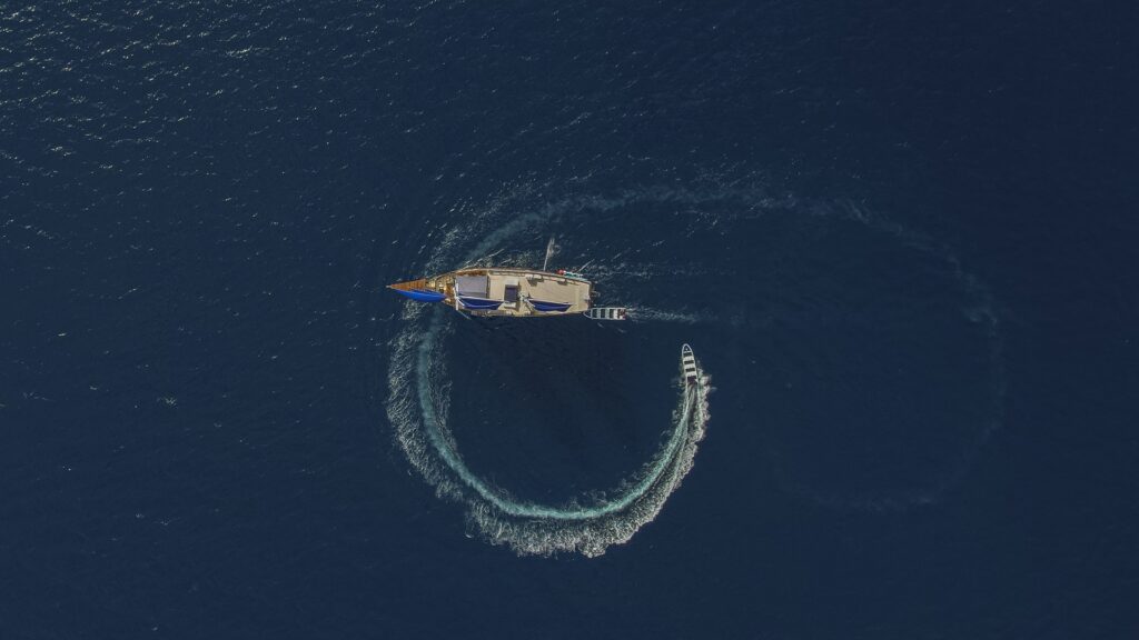 Dramatic overhead drone shot capturing Cajoma V liveaboard surrounded by a sweeping circular wake pattern across open ocean waters.