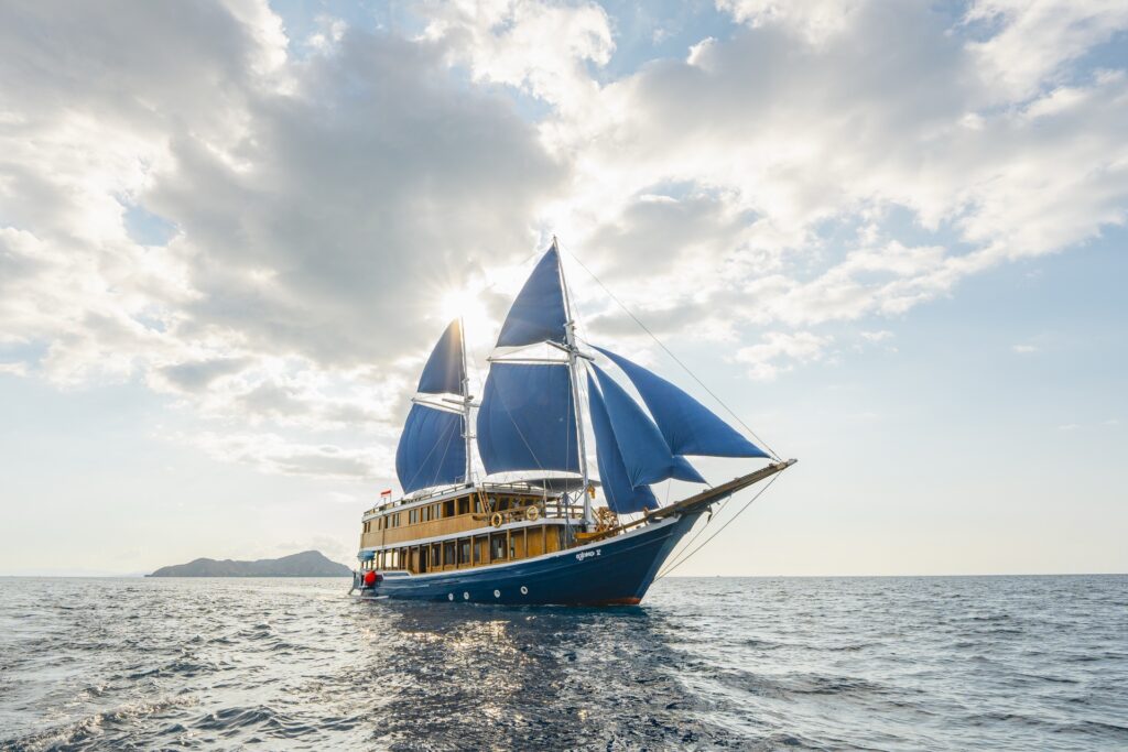 Wide aerial view of Cajoma V luxury yacht anchored peacefully on calm waters with towering blue sails and mountainous island scenery surrounding the vessel.