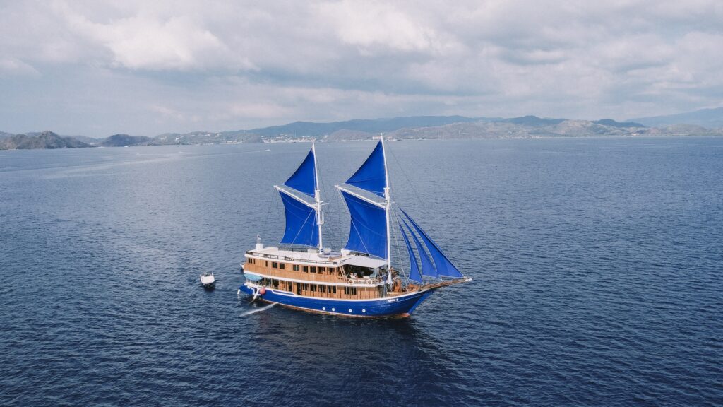 High aerial perspective of Cajoma V luxury yacht gliding through deep blue ocean waters with vibrant blue sails fully extended.