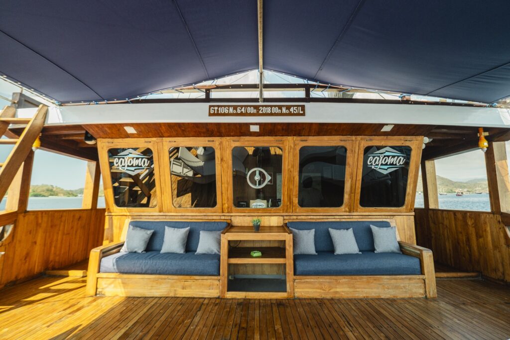 Covered lounge seating area aboard Cajoma V phinisi charter featuring deep blue cushions, warm teak finishes, and shaded open-air relaxation space with panoramic sea views.