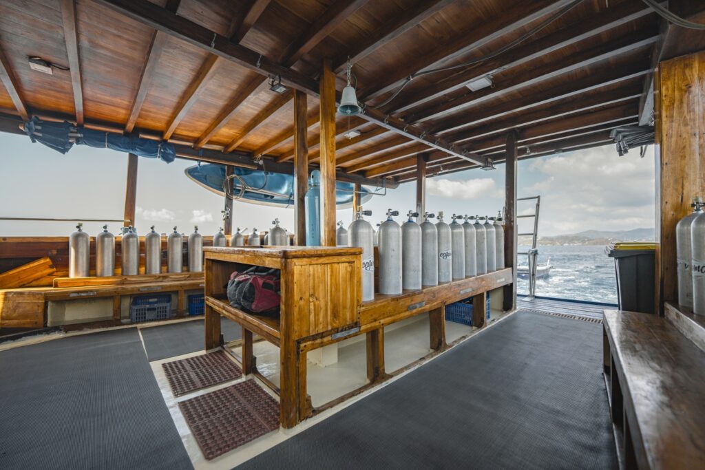 Professional dive deck aboard Cajoma V phinisi charter with neatly arranged scuba tanks, teak equipment benches, and open-air preparation space built for multi-day diving expeditions.
