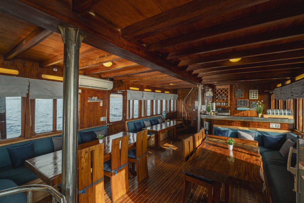 Warm teak dining salon aboard Cajoma V phinisi charter featuring communal tables, cushioned seating, and a cozy onboard bar area designed for social gatherings.