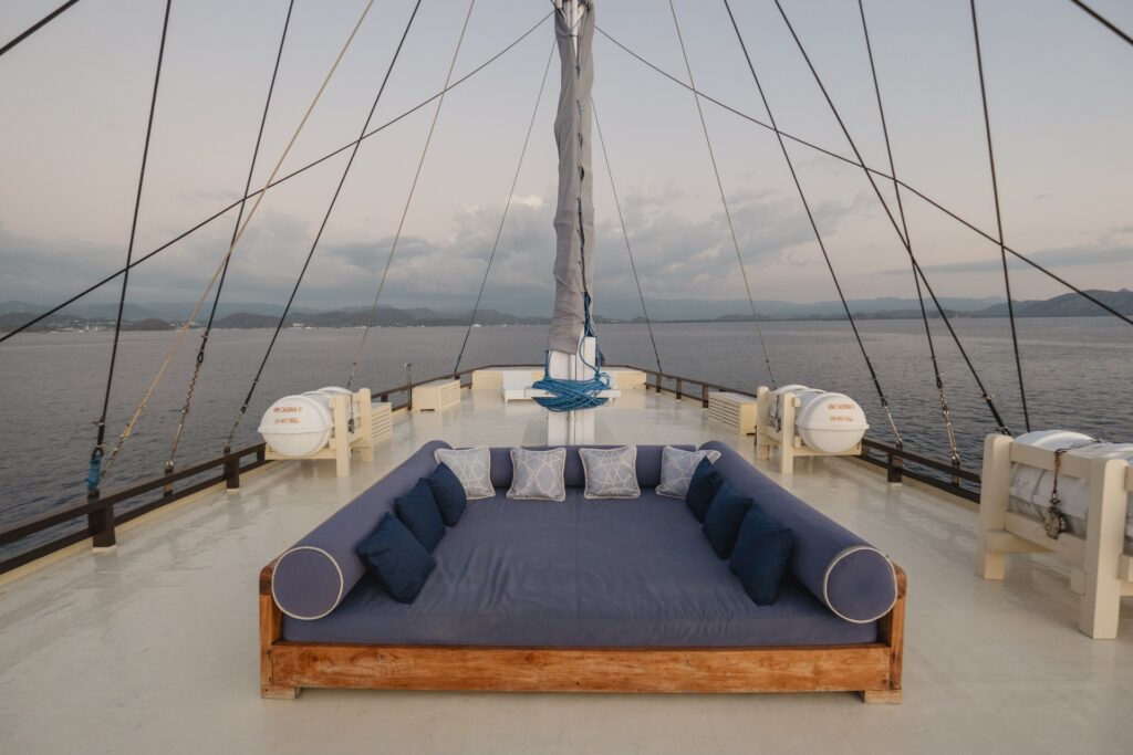 Large rooftop daybed aboard Cajoma V phinisi positioned beneath towering mast lines with tranquil sunset views stretching across the sea.