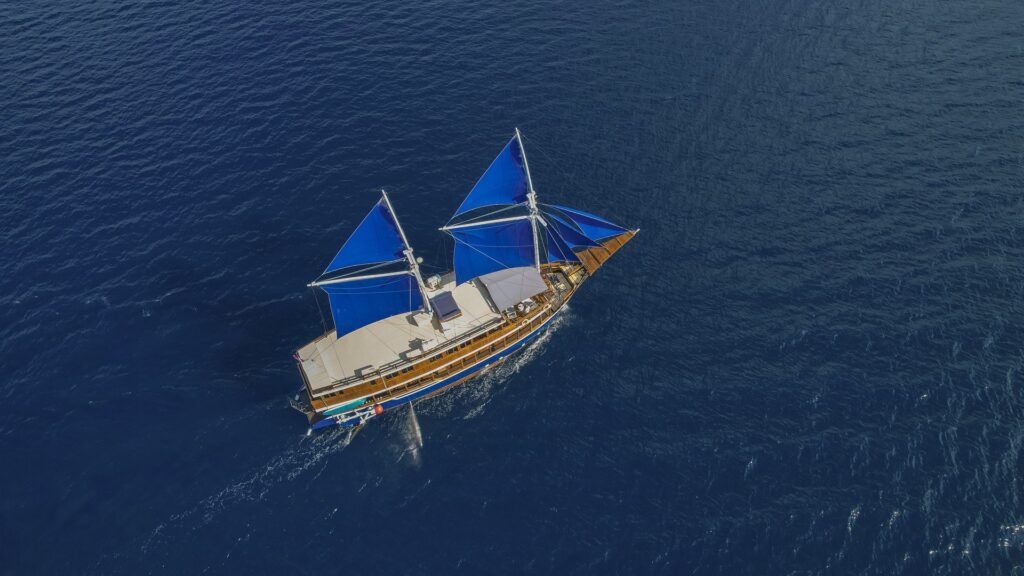 Cajoma V private charter sailing gracefully beneath striking blue sails while golden afternoon light reflects across the calm sea of Komodo Waters.