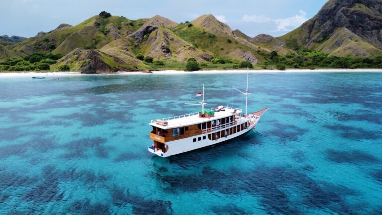 Aerial view of the Dinara phinisi cruising turquoise water near a white sand beach