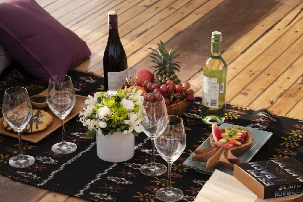 A deck picnic spread of wine, fruit, and appetizers on a woven mat