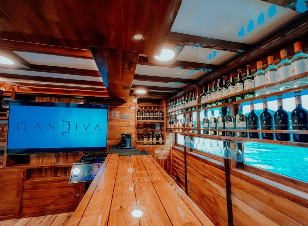 Indoor bar lined with bottle shelves beneath a screen showing the Gandiva logo