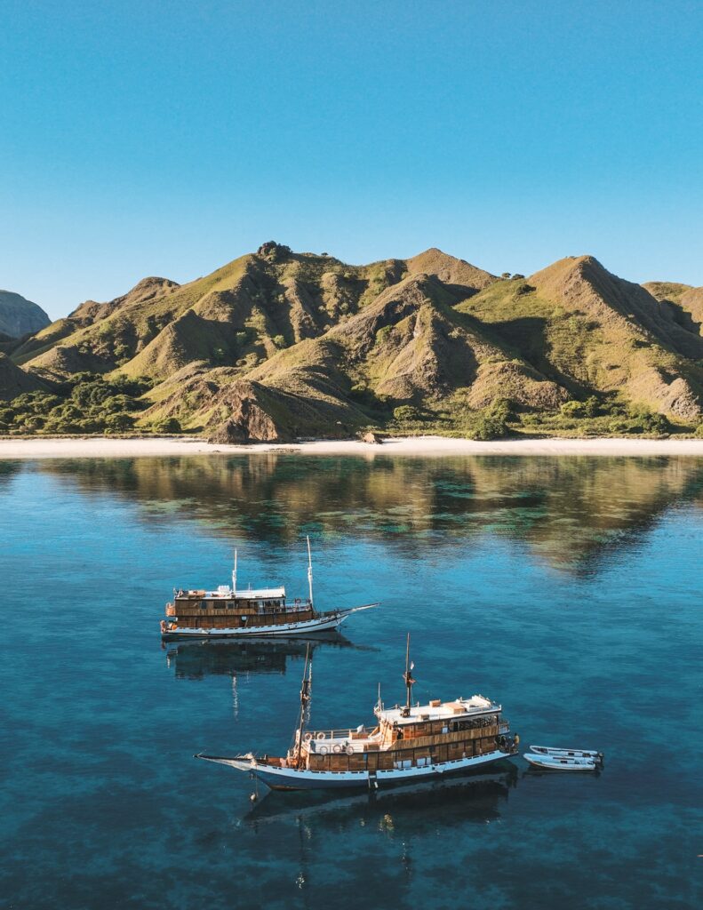 Two liveaboards anchored near a beach and turquoise water below green hills