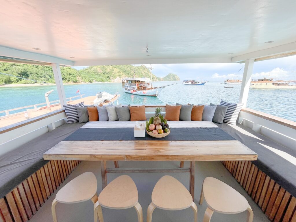 Shaded lounge bench with blue and orange cushions, a wooden table, and white stools overlooking a busy anchorage