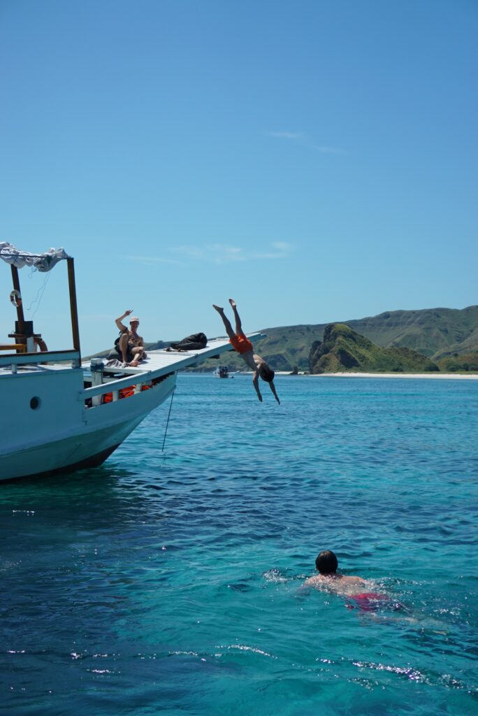 A guest leaping off Zendaya's bow into turquoise water while another swims below near a beach