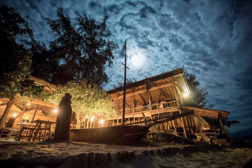 Bamboo beach restaurant lit by lanterns under a full moon, wooden hull on the sand