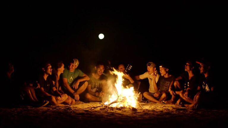 Crew gathered around a beach campfire at night, guitar in hand, full moon above