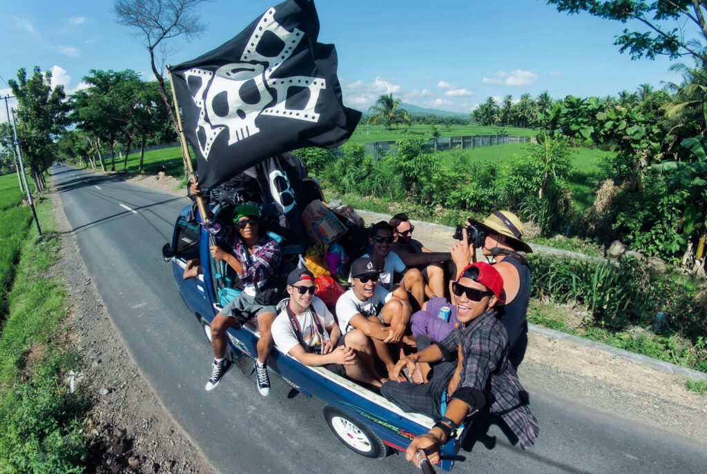 Crew packed into a pickup bed waving a skull flag along a palm-lined country road