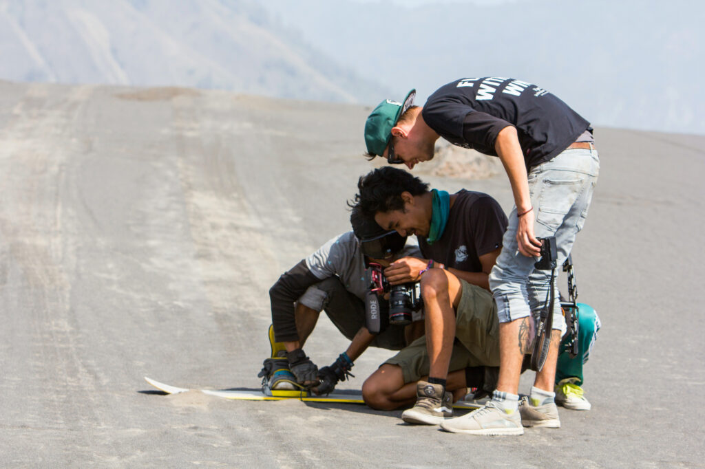 Three film crew members huddle over a camera on a grey volcanic sand plateau