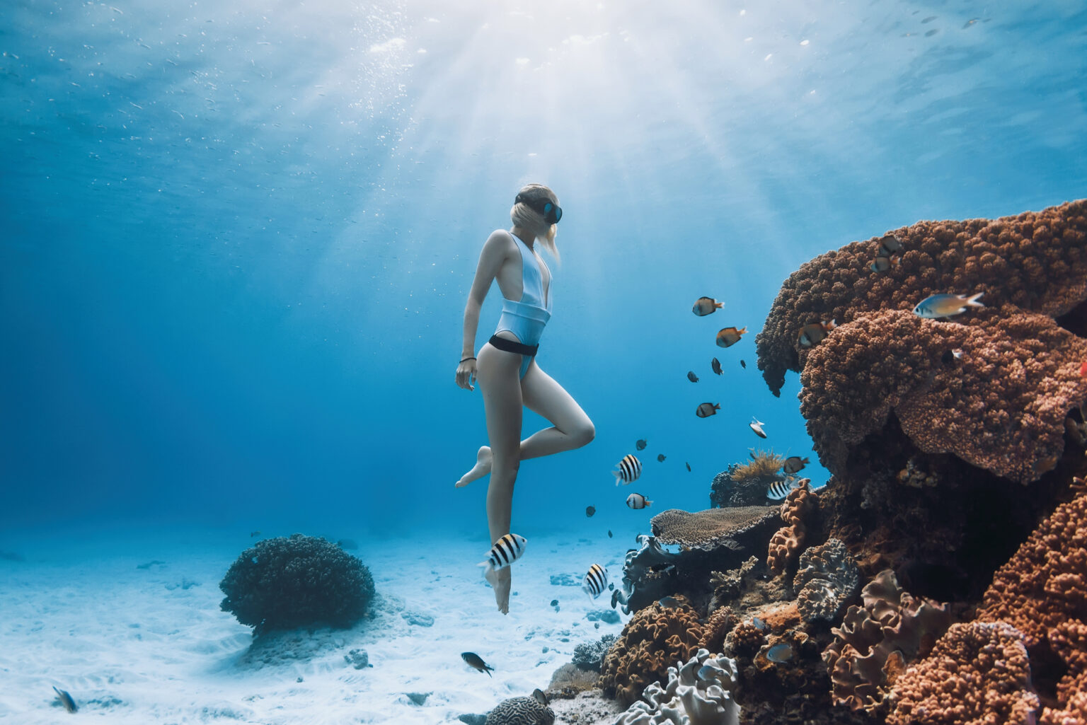 Freediver in a mask hovering beside a coral bommie and reef fish, clear blue water