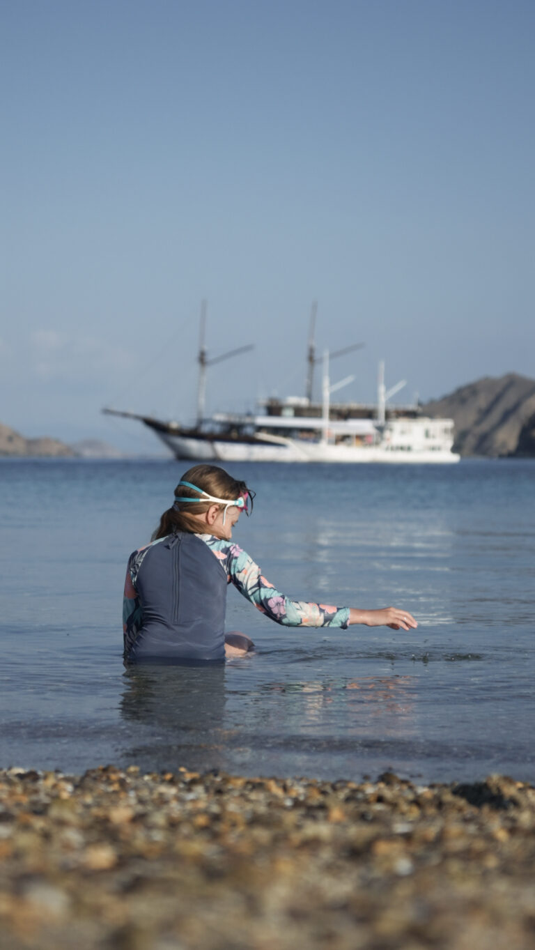 Child in a rash guard sitting in the shallows with a phinisi anchored behind, Komodo