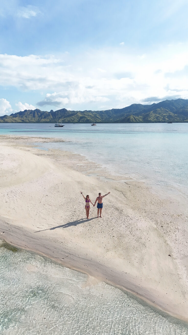 Couple waving from a white sandbar seen from above, green hills behind, Komodo National Park