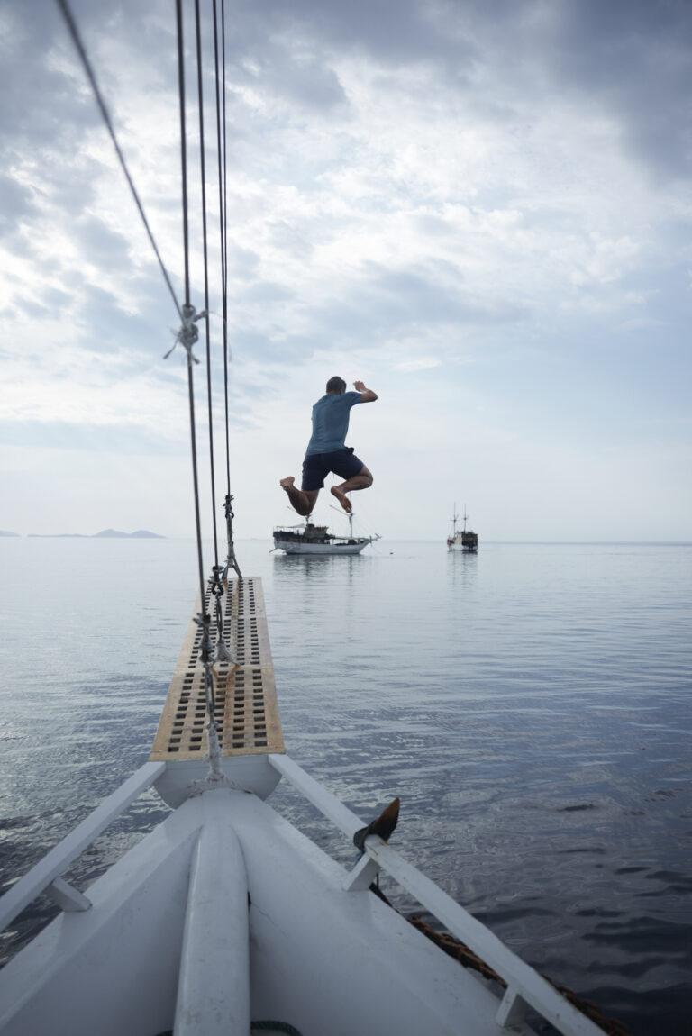 Traveler leaping from the bowsprit of a phinisi into a glassy sea under grey skies