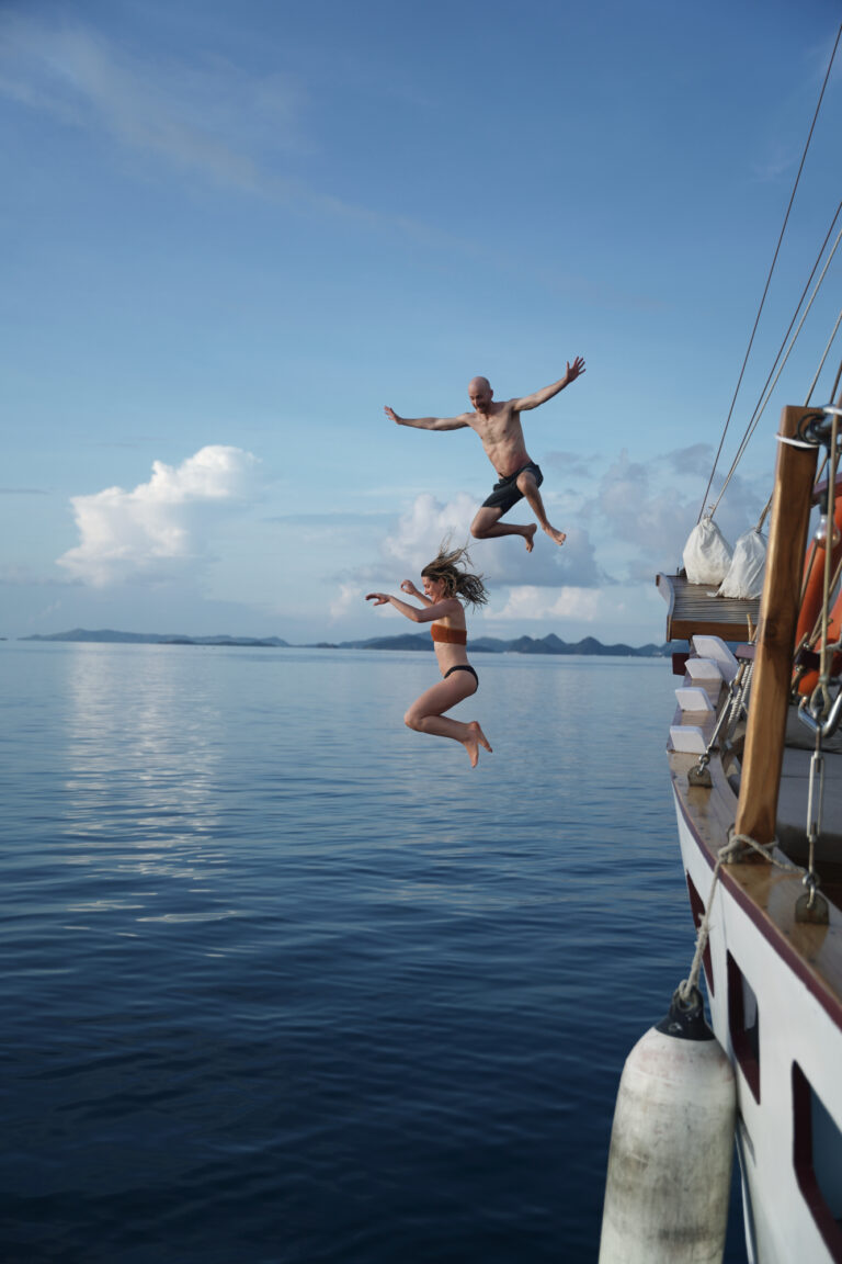 Two travelers mid-air jumping off the side of a phinisi into calm sea, Komodo