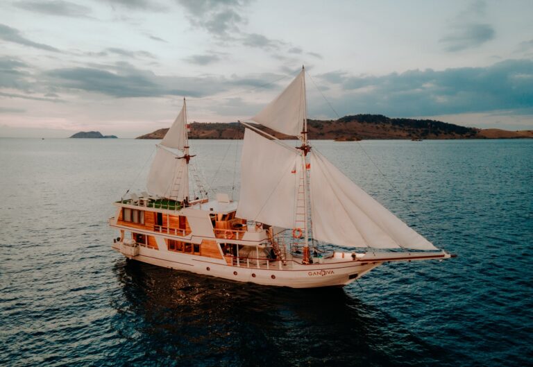 Gandiva phinisi under full sail at dusk, hills silhouetted behind