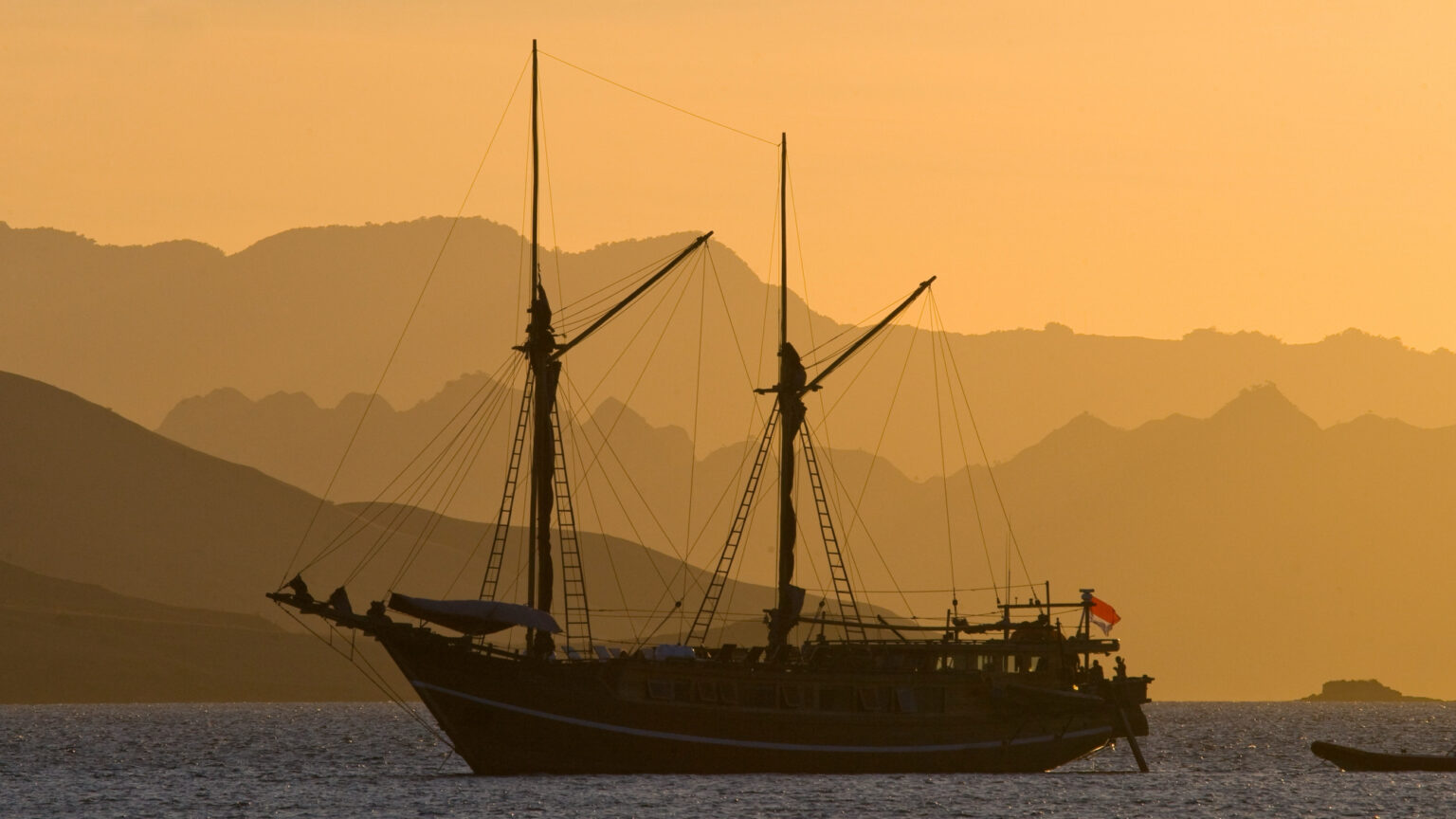 Two-masted Komodo liveaboard phinisi silhouetted at anchor against layered hills at golden sunset