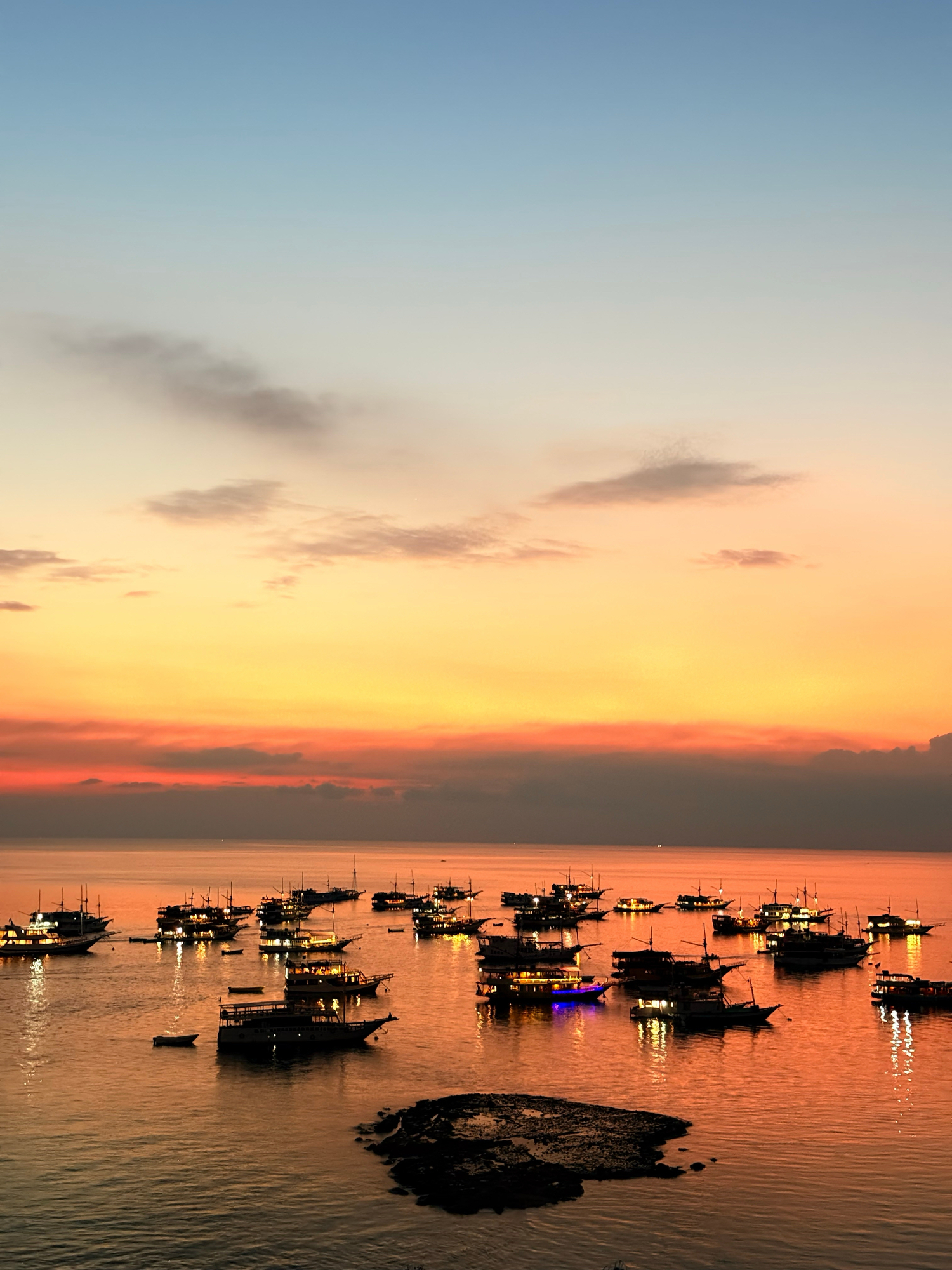 Lit vessels moored on calm water at dusk in Labuan Bajo bay, orange sky above
