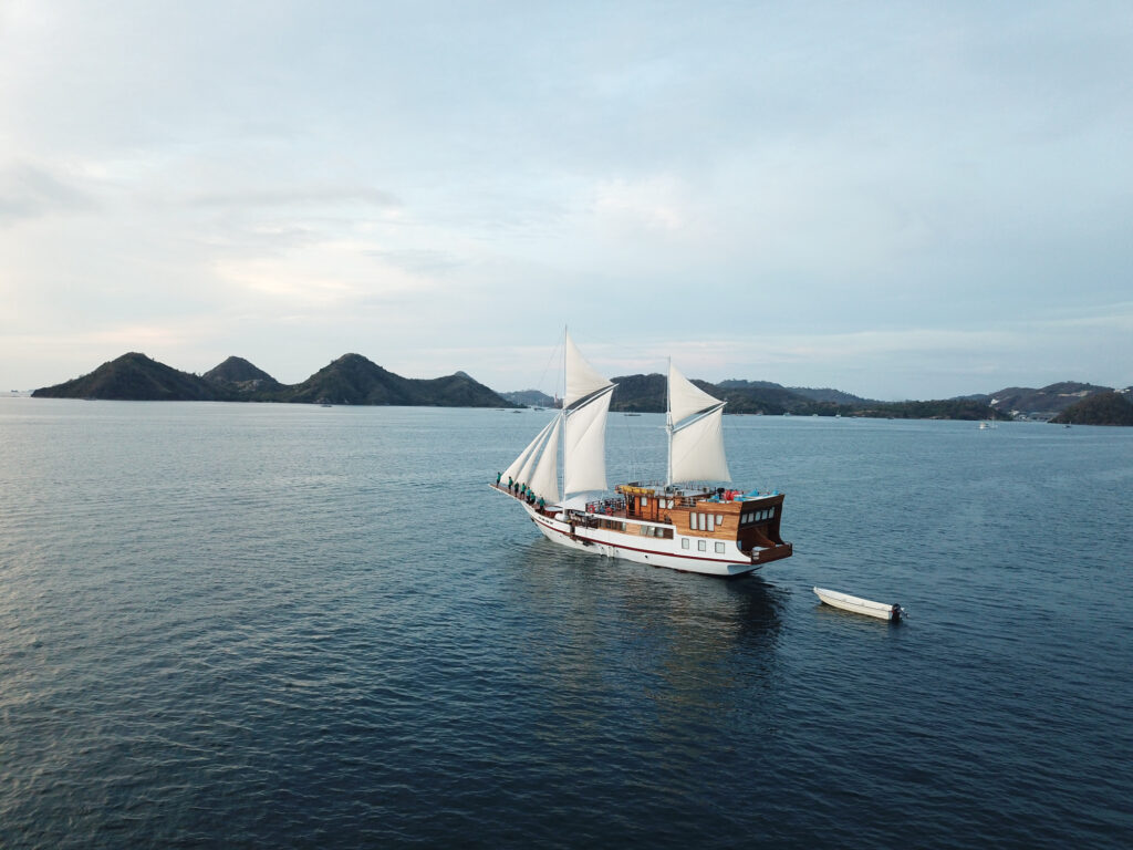 Phinisi under full sail with green hills and a small tender nearby
