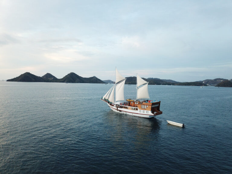 Phinisi under full sail with green hills and a small tender nearby