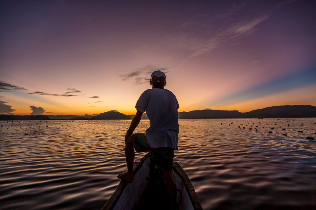 Man seated on the bow of a wooden outrigger canoe at sunset over calm water